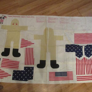 May include: Fabric panel with cut-out shapes for a Liberty Santa doll. The design includes a figure with a yellow body, black boots, and white gloves, along with pieces of the American flag. The text "Fabric Traditions Liberty Santa Doll" is visible.