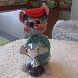 May include: A vintage stuffed mouse toy wearing a red cap and green shirt, holding a silver trophy. The mouse has a painted face with rosy cheeks and a black nose. The trophy has a black base.