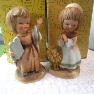 May include: Two porcelain figurines of children dressed in biblical clothing. The figurine on the left is holding a stone tablet. The figurine on the right is holding a small lion. Both figurines are standing on a white lace surface. The figurines are part of a collection called "Little Bible Friends".