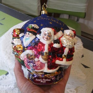 May include: A Christmas ornament featuring multiple Santa Claus figures in red and white suits, with a blue background and gold accents. The ornament has the text "Kringle's Christmas Mingle".