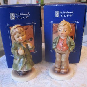 May include: Two porcelain figurines of children in traditional German clothing. The figurines are in their original boxes, which are blue with gold lettering that reads "M.J. Hummel Club".