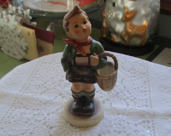May include: A figurine of a boy wearing a green jacket, brown pants, and a red scarf, holding a wicker basket. The figurine is made of porcelain and has a painted finish.