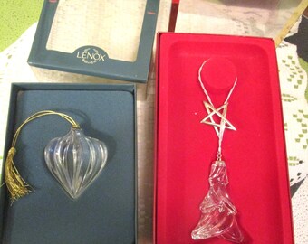 May include: Two Lenox ornaments in their original boxes. One is a clear, ribbed, heart-shaped glass ornament with a gold tassel. The other is a clear, sculpted glass tree ornament topped with a silver-toned star, resting in a red velvet-lined box.