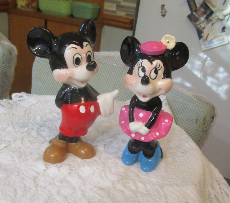 May include: A vintage ceramic figurine set of Mickey Mouse and Minnie Mouse. Mickey is wearing red shorts with white polka dots and brown shoes. Minnie is wearing a pink dress with white polka dots, a pink hat with a white flower, and blue shoes.