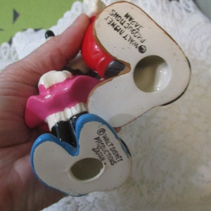 May include: A pair of ceramic figurines of Mickey Mouse and Minnie Mouse. Mickey is wearing red pants and a yellow shirt. Minnie is wearing a pink dress and a white polka dot bow. The figurines are made in Japan and have the Walt Disney Productions logo on the bottom.