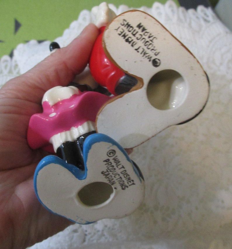 May include: A pair of ceramic figurines of Mickey Mouse and Minnie Mouse. Mickey is wearing red pants and a yellow shirt. Minnie is wearing a pink dress and a white polka dot bow. The figurines are made in Japan and have the Walt Disney Productions logo on the bottom.