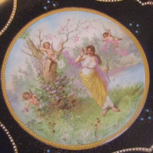 May include: A vintage porcelain plate with a black background and a gold rim. The center features a colorful scene of a woman with a bow and arrow, two cherubs, and a blooming tree. The plate is decorated with gold and blue accents.