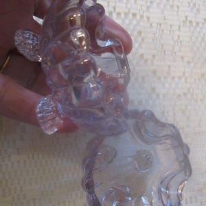 May include: A clear, decorative glass ashtray with a unique, swirling design. The ashtray has a circular bowl shape with a scalloped edge and is held by a hand. The glass has a slight purple hue.