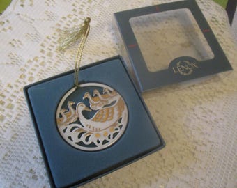 May include: A circular white porcelain ornament featuring gold-painted swan motifs and cut-out details. The ornament includes a gold hanging cord and is presented in a blue box with a clear lid, labeled with the brand name Lenox in gold lettering.