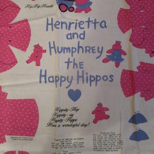 May include: A white fabric with pink hippopotamus illustrations and the text "Henrietta and Humphrey the Happy Hippos." The design includes a heart, text, and instructions for sewing the hippos.