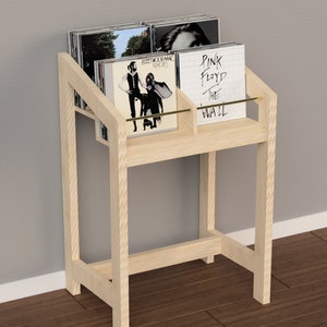 Puede incluir: Un soporte para discos de madera clara con dos estantes y una barra de metal dorado. El soporte contiene discos de vinilo con portadas de álbumes en blanco y negro. Las portadas de los álbumes incluyen "Pink Floyd The Wall" y "New York Dolls".