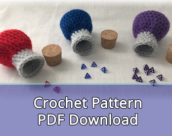 Peut inclure: Trois fioles de potion crochetées avec des bouchons en liège, une rouge, une bleue et une violette. Chaque fiole est remplie de petits dés violets transparents.  Modèle de crochet Téléchargement PDF.