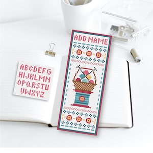 May include: A bookmark with a cross-stitch design of a basket of yarn and knitting needles. The bookmark is white with red, blue, and yellow accents. The text "ADD NAME" is at the top of the bookmark.