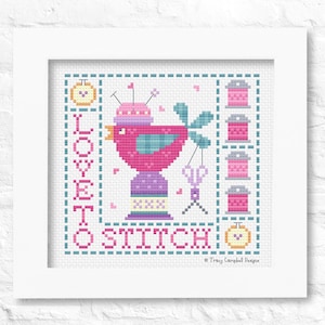 May include: Cross-stitch artwork in a white frame. The design features a pink bird with a sewing theme, including spools of thread, scissors, and the text "LOVE TO STITCH". The artwork is in a square format.