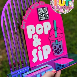 Puede incluir: Un letrero acrílico multicolor con las palabras "Pop & Sip" y una ilustración de té de burbujas. El letrero tiene un código QR y el texto "Get Social With Us". El letrero está sostenido por una mano.