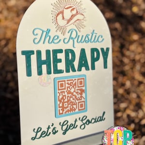 May include: A white sign with a rustic cowboy hat design and the text "The Rustic Therapy" in blue and green lettering. The sign also includes a QR code and the text "Let's Get Social" in green lettering.