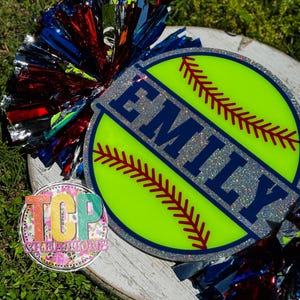 May include: A softball-themed decoration with the name "EMILY" in navy blue, set against a neon green background. The design includes red stitching details and a silver glitter border. Colorful pom-poms in red, blue, and silver add a festive touch.