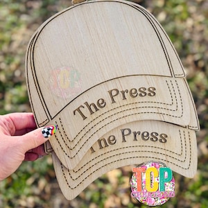 Puede incluir: Tres gorras de béisbol de madera apiladas una encima de la otra. La gorra superior tiene las palabras "The Press" grabadas en la parte delantera. La gorra del medio tiene las palabras "The Press" grabadas en la parte delantera. La gorra inferior tiene un logotipo en la parte delantera.