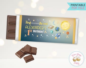 May include: Printable chocolate bar wrapper with a space theme. It features a yellow sun, planets, stars, and a rocket. The text reads "First trip around the sun ALEXANDER'S 1ST Birthday!"