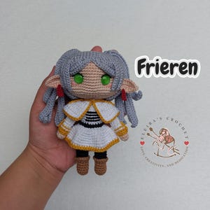 May include: A crocheted doll with grey hair, green eyes, and a white and yellow dress. The doll is holding a red and white striped scarf. The doll is labelled "Frieren".
