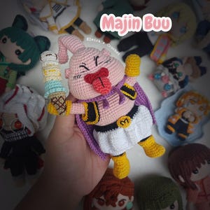 May include: A handmade crochet doll of Majin Buu from Dragon Ball Z, holding a colorful ice cream cone. The doll is pink, with a purple cape and yellow boots. Other anime-style crochet dolls surround it.