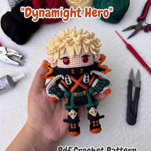 May include: A crocheted doll with blonde hair and red eyes, dressed in a black and green outfit with orange details. The doll is held in a hand, with the text "Dynamight Hero" and "Pdf Crochet Pattern" visible.