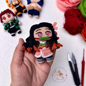 May include: Crocheted anime character dolls, featuring Nezuko in a pink kimono, held in a hand. Other characters include Tanjiro, Zenitsu, and Inosuke. Yarn and crochet tools are visible in the background. The dolls are handmade.