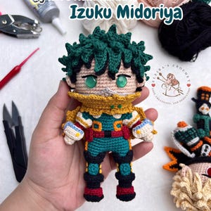 May include: A crocheted amigurumi doll of Izuku Midoriya from My Hero Academia, featuring spiky dark green hair, green eyes, a yellow collar, and a teal, red, and black suit. The text My Hero Academia's Izuku Midoriya and Pdf Crochet Pattern appear.