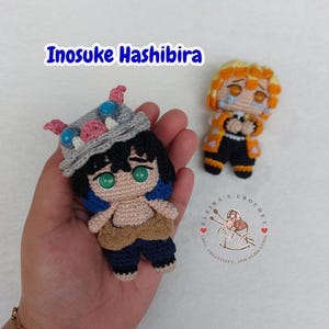 Op de afbeelding: Gehaakte Inosuke Hashibira en Zenitsu Agatsuma figuren. De Inosuke figuur wordt in een hand gehouden en heeft een grijze everzwijnkop, zwart haar en blauwe en roze accenten. De Zenitsu figuur is geel en oranje.