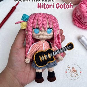 Puede incluir: Una muñeca de ganchillo de Hitori Gotoh de Bocchi The Rock! con pelo rosa, accesorios azules y amarillos, chaqueta color melocotón, falda gris, calcetines negros y zapatos marrones. La muñeca sostiene una guitarra negra con detalles dorados.