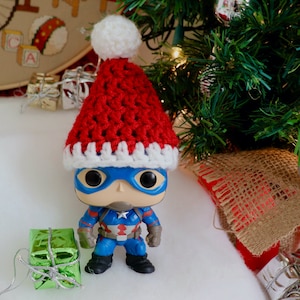 May include: A red and white crocheted Santa hat sits on top of a blue and red Captain America action figure. The figure is wearing a blue and red suit with a white star on the chest. The figure is standing on a white surface.