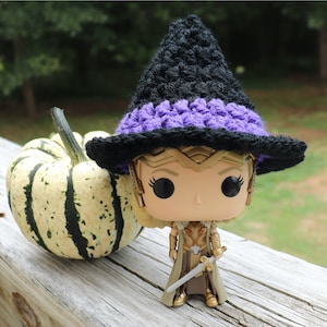 May include: A black and purple crocheted witch hat with a wide brim. The hat is sitting on top of a small, golden figurine with a sword.