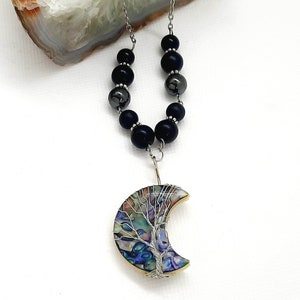 May include: A silver chain necklace with black beads and a crescent moon pendant featuring a wire-wrapped tree of life design. The pendant is made of iridescent abalone shell.