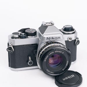May include: A silver and black Nikon film camera with a Nikon Lens Series E 50mm 1:1.8 lens attached. The camera body features a textured black grip, a silver top plate with various dials, and a hot shoe. A separate black Nikon lens cap sits nearby.