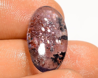 May include: Oval-shaped gemstone with a translucent, purplish-pink hue. The stone features sparkling inclusions of various sizes and colors, including red, silver, and black, creating a unique, glittery effect. The gemstone is held between fingers.
