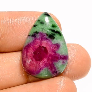 Pode incluir: Uma pedra preciosa em forma de gota com uma superfície polida. A pedra apresenta uma vibrante mistura de cores, incluindo uma área central de vermelho profundo e uma matriz verde circundante com inclusões pretas. A gema é segurada em uma mão.