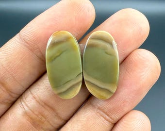 May include: Two oval-shaped gemstones with a polished finish. The stones feature a blend of olive green, tan, and cream colors, with natural banding patterns. The gemstones are held in a hand.