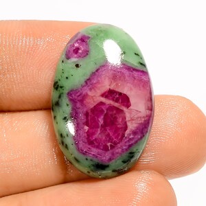 May include: An oval gemstone with a striking colour scheme. The stone has a central area of deep red, encircled by a lighter pink shade, and set within a green matrix dotted with black. The polished surface reflects light.