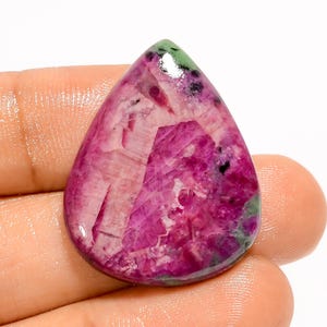 Könnte beinhalten: Ein tropfenförmiger, polierter Edelstein. Der Stein zeigt eine Mischung aus Rosa- und Magentatönen mit grünen und schwarzen Bereichen. Der Edelstein wird in einer Hand gehalten, um seine Form und Größe zu zeigen.