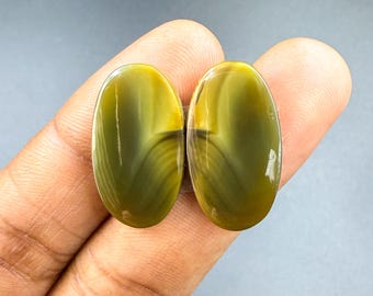 May include: Two oval-shaped gemstones with a polished finish. The stones have a color gradient of olive green to yellow, with dark bands. The gemstones are held in a hand against a gray background.