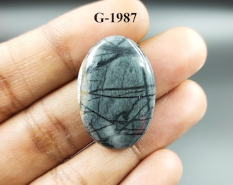 May include: Oval cabochon gemstone with a gray and black pattern. The stone has a smooth, polished surface. G-1987