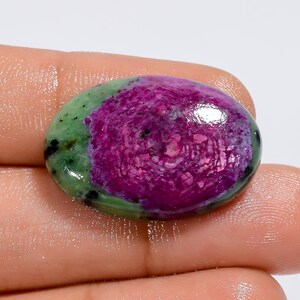 Puede incluir: Gema ovalada con una vibrante combinación de colores. El centro presenta un tono magenta intenso, rodeado por un borde verde con motas negras. La superficie pulida refleja la luz, mostrando la belleza natural de la piedra.