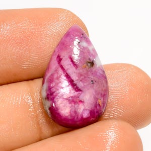 Puede incluir: Una gema en forma de lágrima de color rosa vibrante, con patrones internos de rosa más oscuro y blanco. La superficie pulida refleja la luz, mostrando la belleza natural de la piedra. La gema se sostiene entre los dedos.