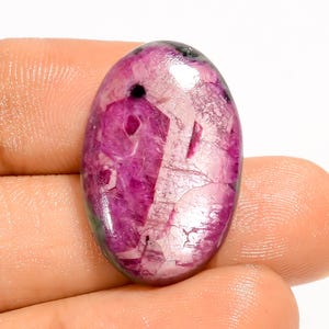 Puede incluir: Una gema ovalada de color rosa vibrante, con una mezcla de tonos más claros y oscuros. La piedra tiene una superficie pulida con patrones e inclusiones internas visibles, y es sostenida por una mano.