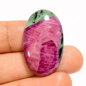 May include: An oval-shaped gemstone with a polished surface. The stone features a vibrant mix of colors, including deep pink, ruby red, green, and black. The gemstone is held in a hand, showcasing its size and shape. The stone is likely a ruby.