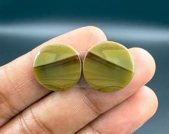 May include: Two round, polished gemstones with a layered design. The stones feature a mix of olive green and brown hues, with a diagonal stripe across each. The gemstones are held in a hand, showcasing their smooth, circular shape.