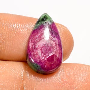 May include: A polished, teardrop-shaped gemstone. The stone displays a mix of colours: deep red, pink, green, and black. The gemstone is held between fingers, highlighting its size and form.