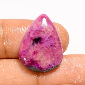 Può includere: Una gemma lucida a forma di goccia, con una vibrante miscela di tonalità rosa e viola. La pietra presenta un aspetto marmorizzato con inclusioni più scure, quasi nere. La base della pietra ha un colore grigio-verde più scuro.