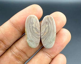 May include: Two oval-shaped gemstones with a swirling, layered pattern in shades of beige, gray, and hints of green. The stones are held in a hand against a dark background. These stones are suitable for jewelry making.