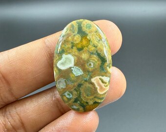May include: Oval-shaped gemstone with a polished surface. The stone features a pattern of green, yellow, and brown swirls and spots. The gemstone is held in a hand against a dark background. The stone is approximately 2 inches long.